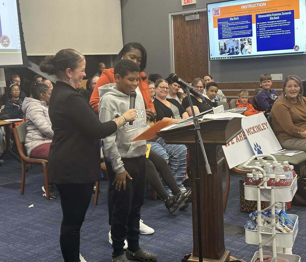 A male student is speaking into a microphone, held by a district staff member, at a podium in a conference room. There is an audience of people in the background looking attentively. District staff members are standing close to the student. A presentation is being displayed in the background.