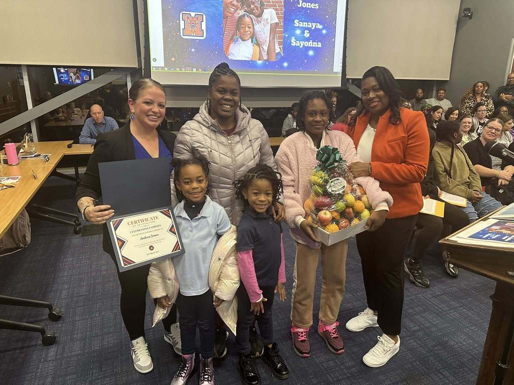 District staff members, one adult and three children are standing in a conference room. One of the district staff members is holding a certificate in her hand and one of the young girls is holding a fruit basket in her hand. A presentation is being displayed in the background. People can be seen sitting in chairs and standing in the background.