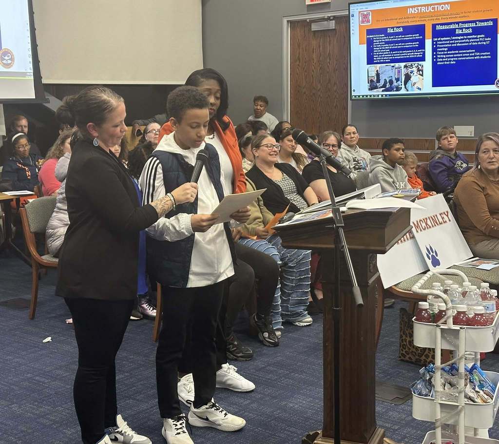 A male student is speaking into a microphone, held by a district staff member, at a podium in a conference room. There is an audience of people in the background looking attentively. District staff members are standing close to the student. A presentation is being displayed in the background.
