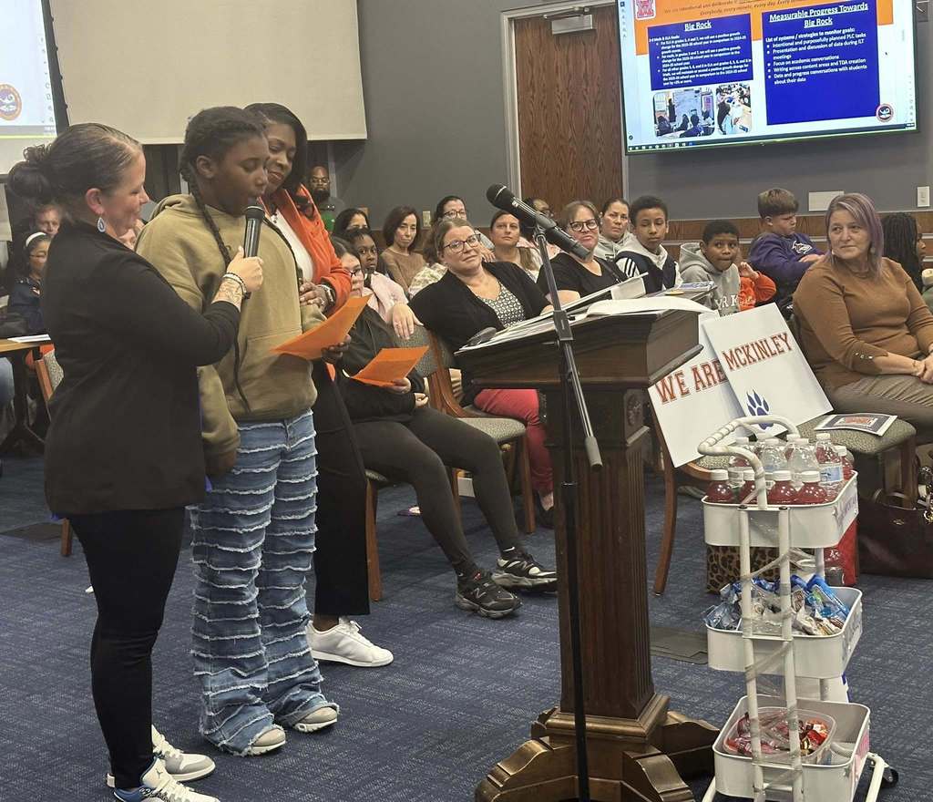 A female student is speaking into a microphone, held by a district staff member, at a podium in a conference room. There is an audience of people in the background looking attentively. District staff members are standing close to the student. A presentation is being displayed in the background.