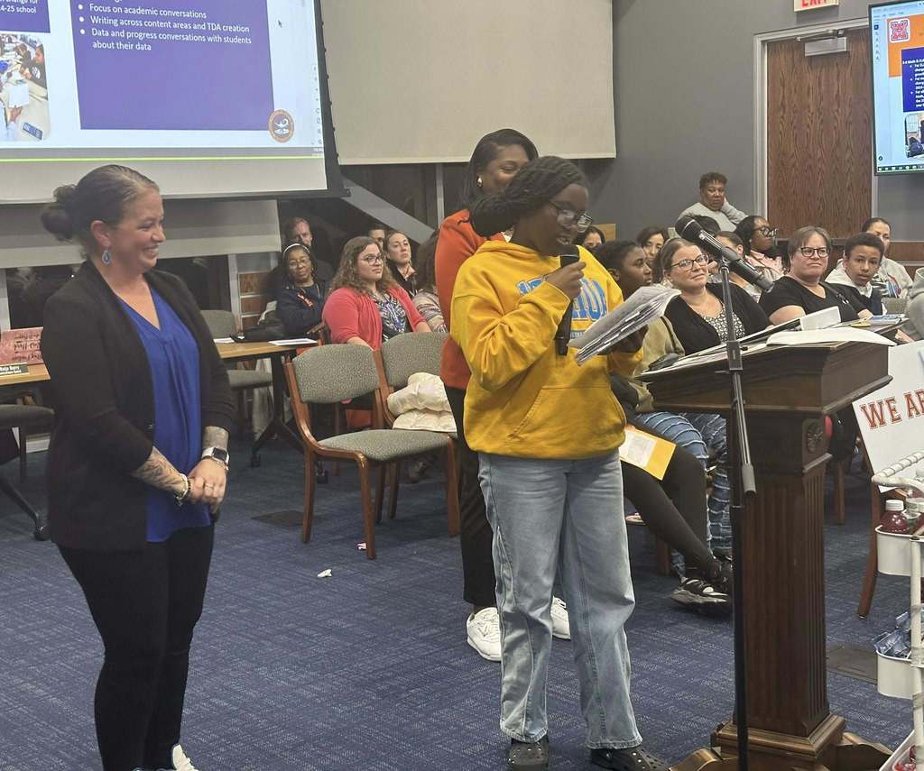 A female student is speaking into a microphone at a podium in a conference room. There is an audience of people in the background looking attentively. District staff members are standing close to the student. A presentation is being displayed in the background.