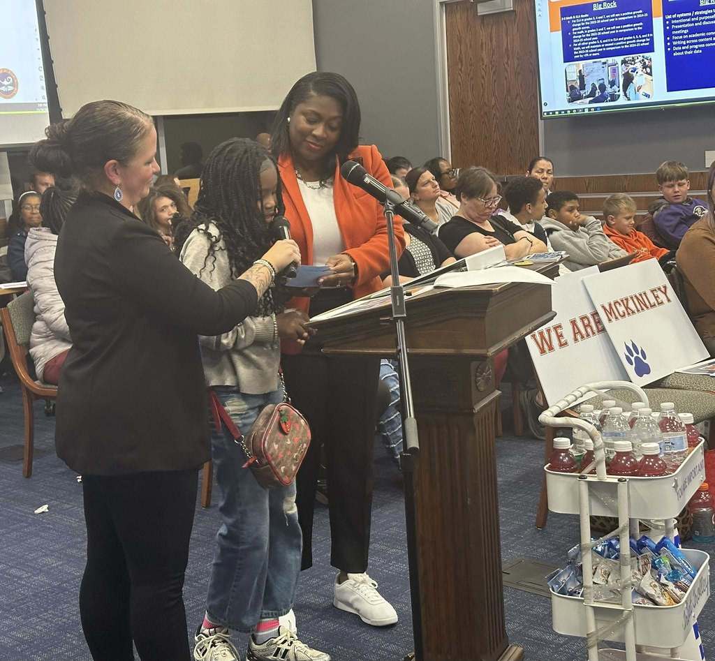 A female student is speaking into a microphone, held by a district staff member, at a podium in a conference room. There is an audience of people in the background looking attentively. District staff members are standing close to the student. A presentation is being displayed in the background.