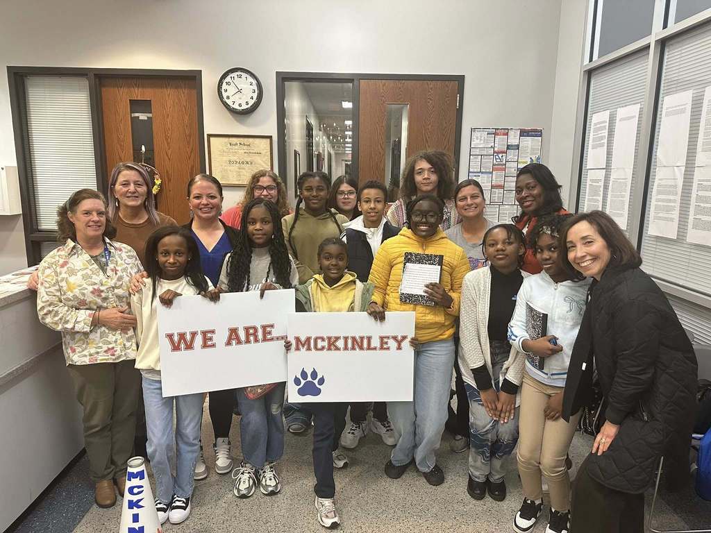 A group of students and district staff members are standing in a lobby area. Four of the students are holding up signs that say  "We Are McKinley" and two of the students are holding composition notebooks in their hands.