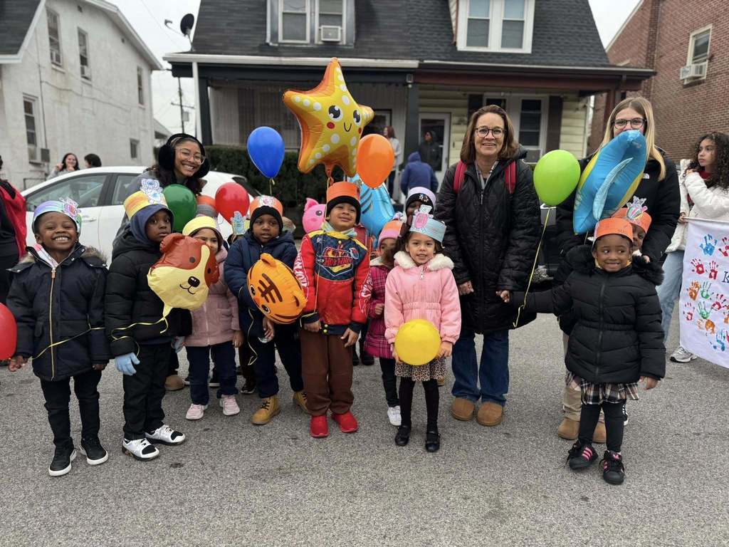 A group of district staff members and students are standing in the middle of a street holding colorful balloons that are shaped like stars and animals. 