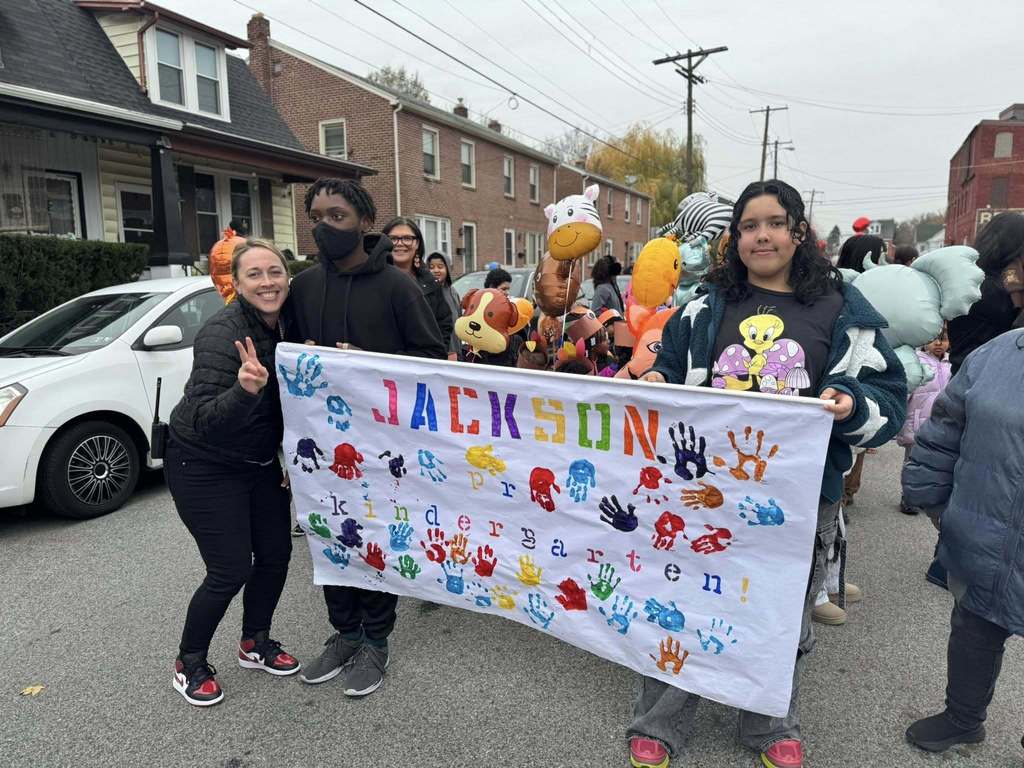 A district staff member and a group of students are standing in the middle of a street holding a colorful banner that says "Jackson Kindergarten" and is covered with multicolored handprints. 
