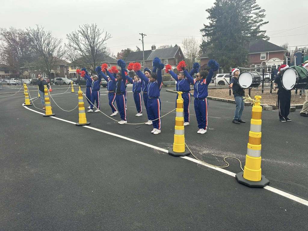 The York High Bearcat Varsity Cheerleaders are holding orange and blue pom-poms in the air, while standing next to members of the William Penn Marching Band who are holding drums in their hands. Yellow cones are lining the road. 