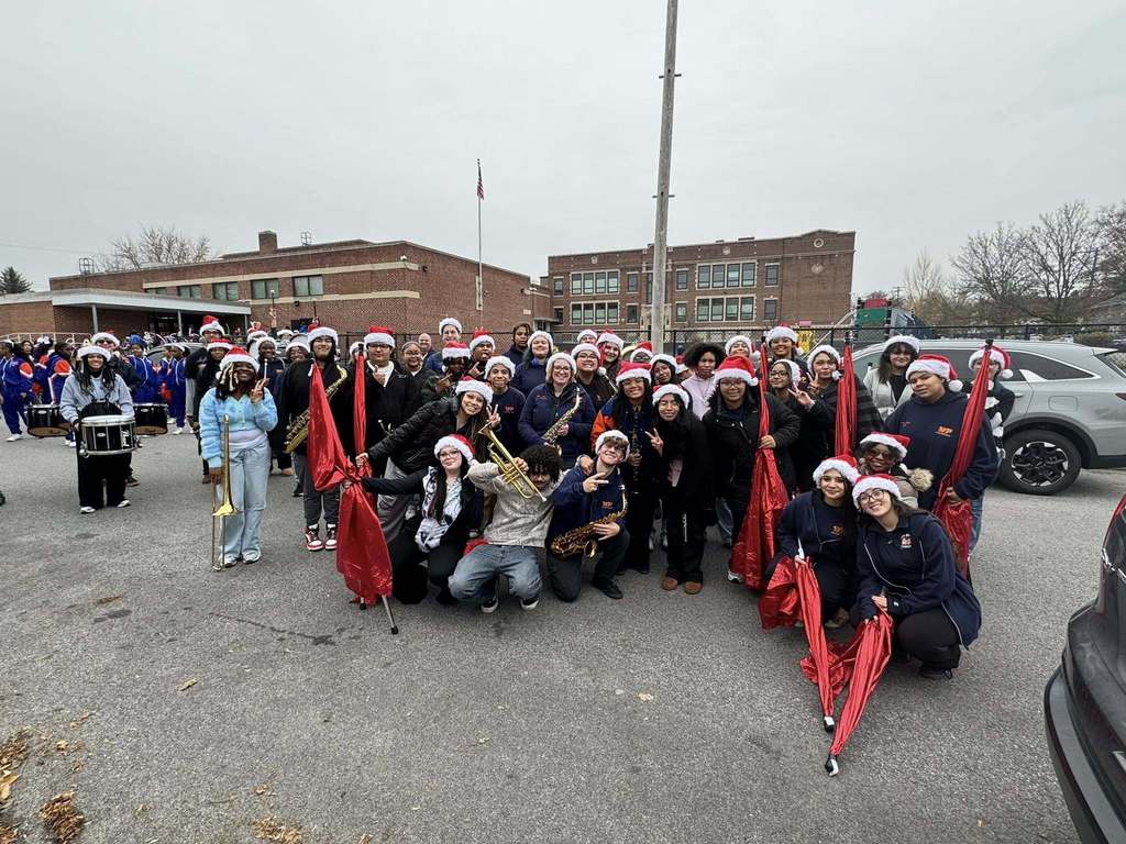 A group of students and district staff members who are apart of the William Penn Marching Band are standing outside wearing Santa hats and holding musical instruments and red flags. 