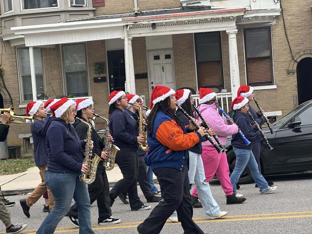 The William Penn Marching Band are playing instruments, including saxophones and clarinets, while wearing Santa hats. They are walking down a residential street.
