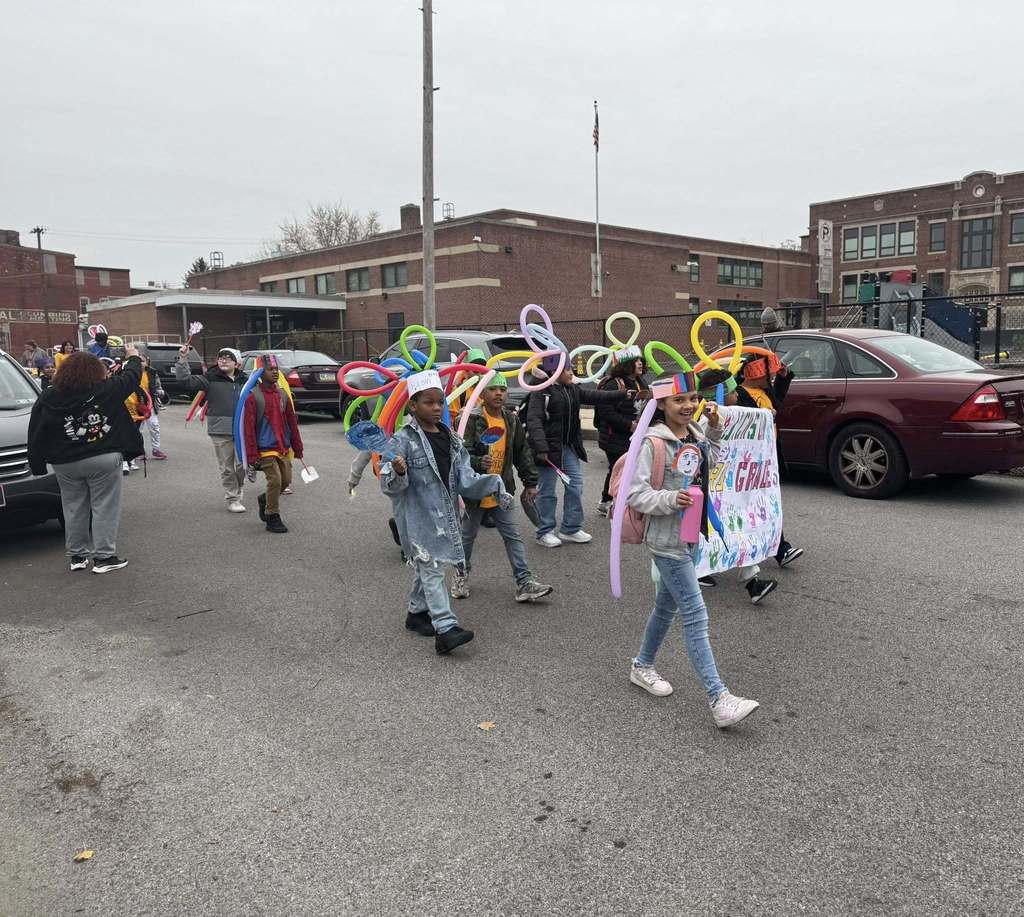 A group of students are parading down a street, wearing colorful balloon hats. Some of the students are holding a decorated sign.