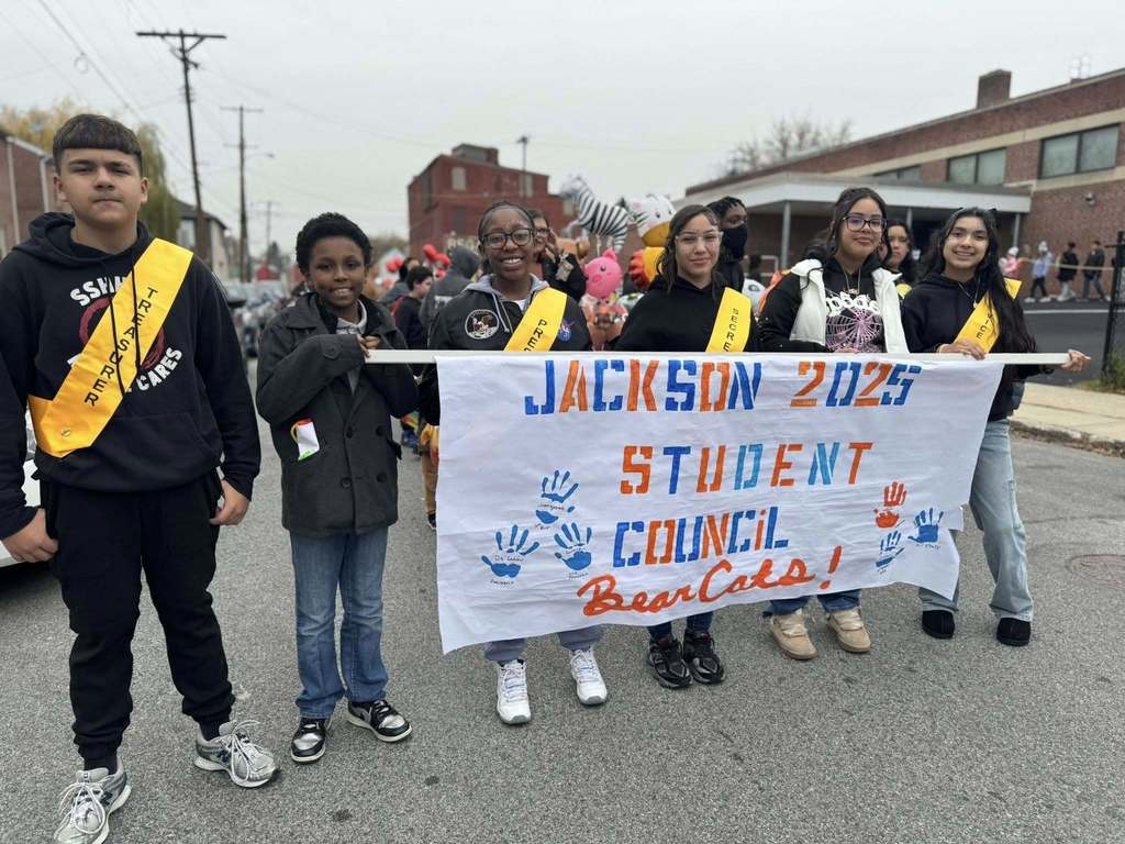 A group of six students are standing in the middle of a street holding a "Jackson 2025 Student Council BearCats" banner. Some of the students are wearing yellow slashes.