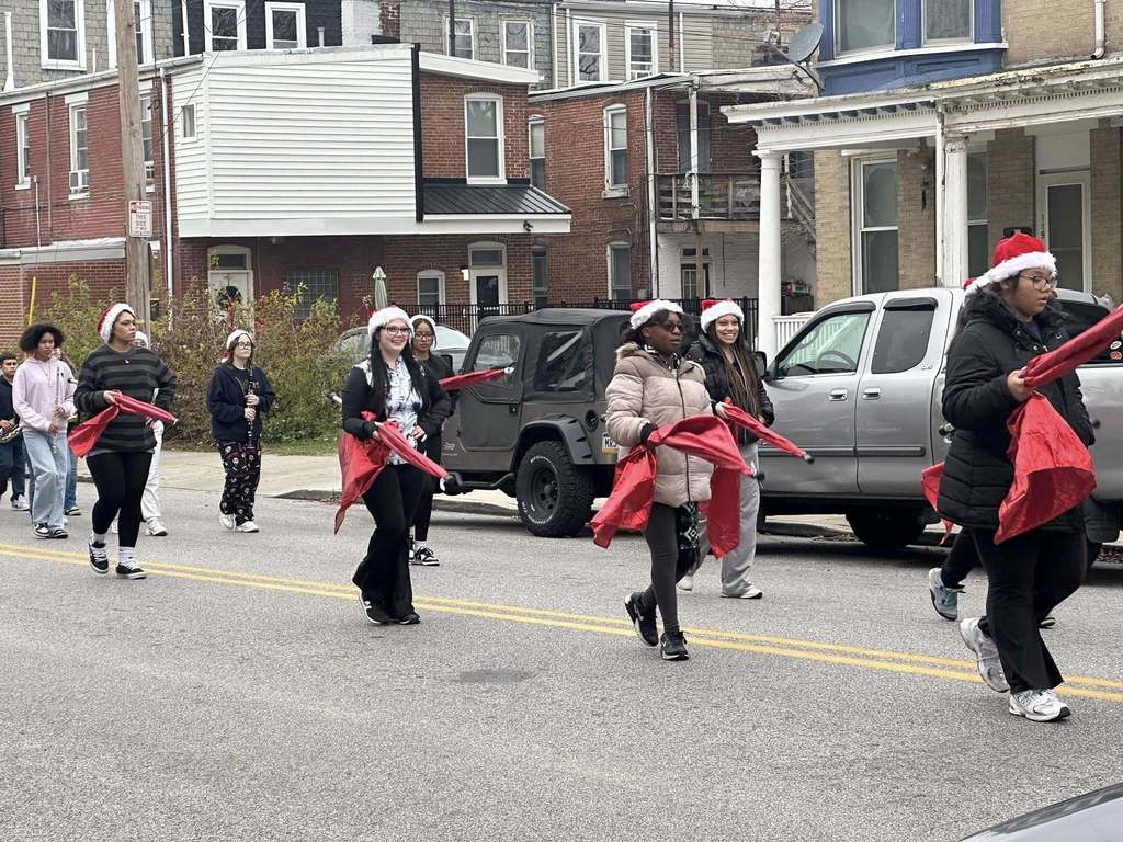 A group of students from the William Penn Marching Band are wearing Santa hats and carrying red flags while walking down a street in a parade.