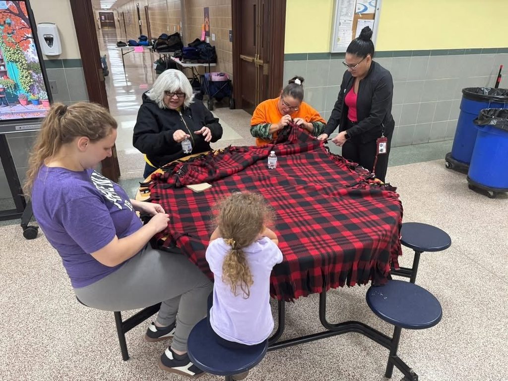 A group of four adults and one student are sitting around a table in a school cafeteria tying knots in a black-and-red plaid fabric.