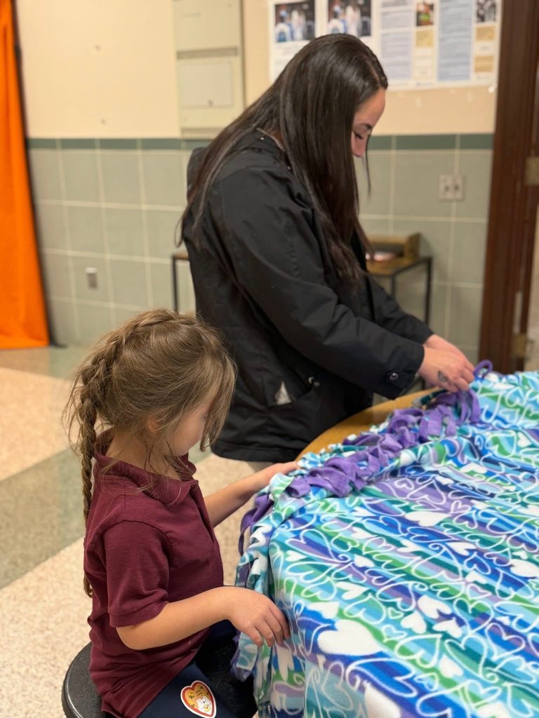 An adult and a child are collaborating on a colorful fabric project in a school cafeteria.