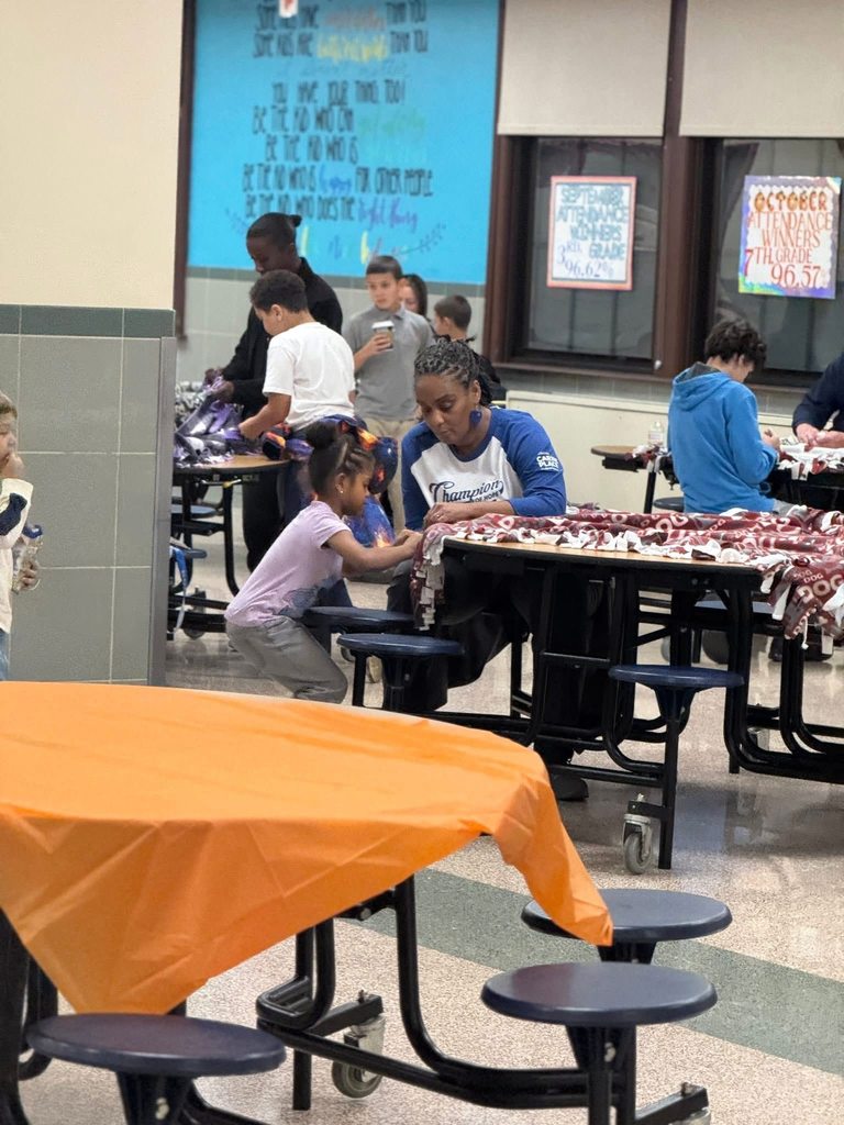 A group of children and district staff members are engaging in crafting activities in a school cafeteria. The district staff member is helping a young girl tie fabric, while vibrant posters are hanging nearby on the walls.
