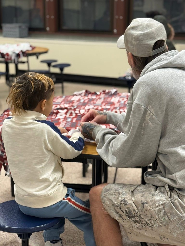 An adult and a child are sitting at a table working on a colorful fabric project in a school cafeteria.