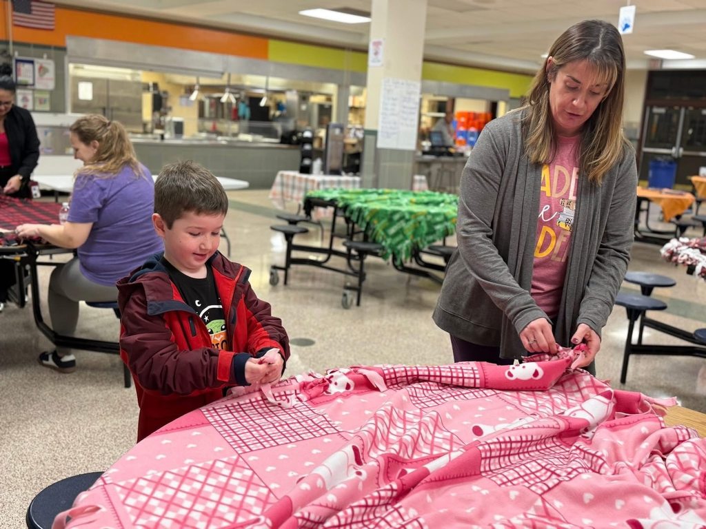 A district staff member and a young boy are making a fleece blanket at a table in a school cafeteria, while other people are in the background working on crafts.
