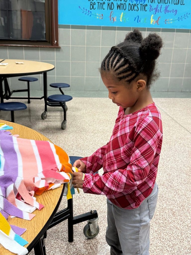 A student is standing near a table in a school cafeteria holding a piece of a colorful blanket. 