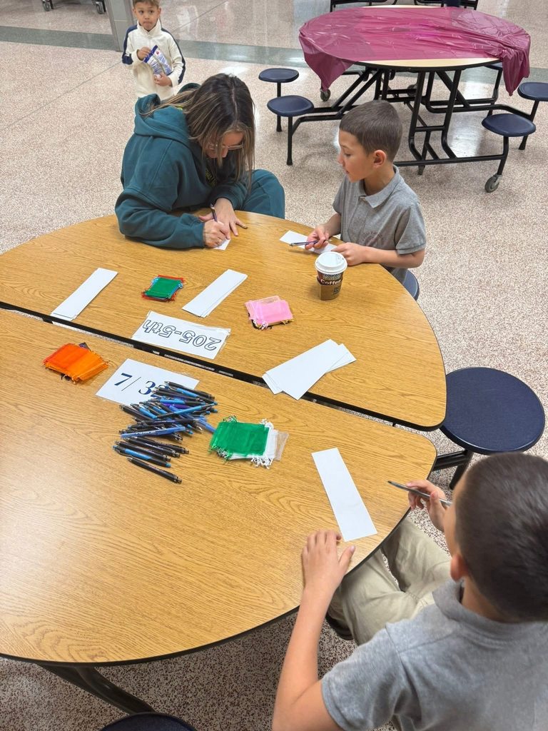 An adult woman and two young boys are sitting around a table in a school cafeteria, writing on papers. There are pens, colored fabric squares, and a coffee cup that are on the table.