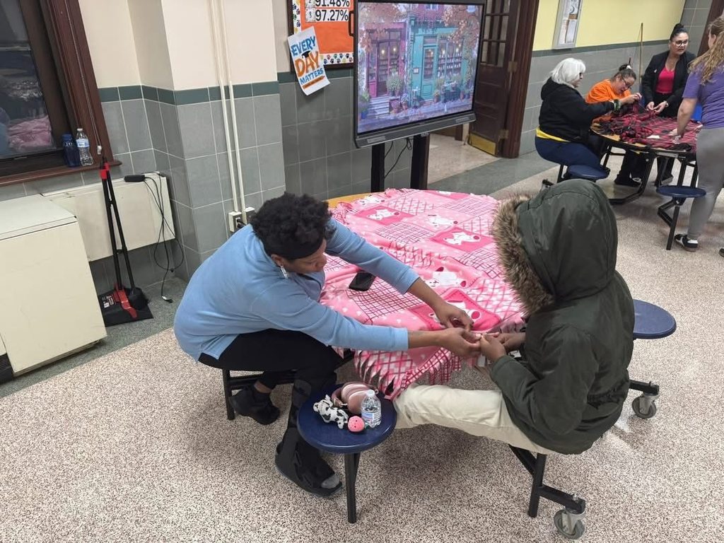 An adult is assisting a young person with a crafting activity in a school cafeteria. Other people can be seen working on a craft activity in the background.