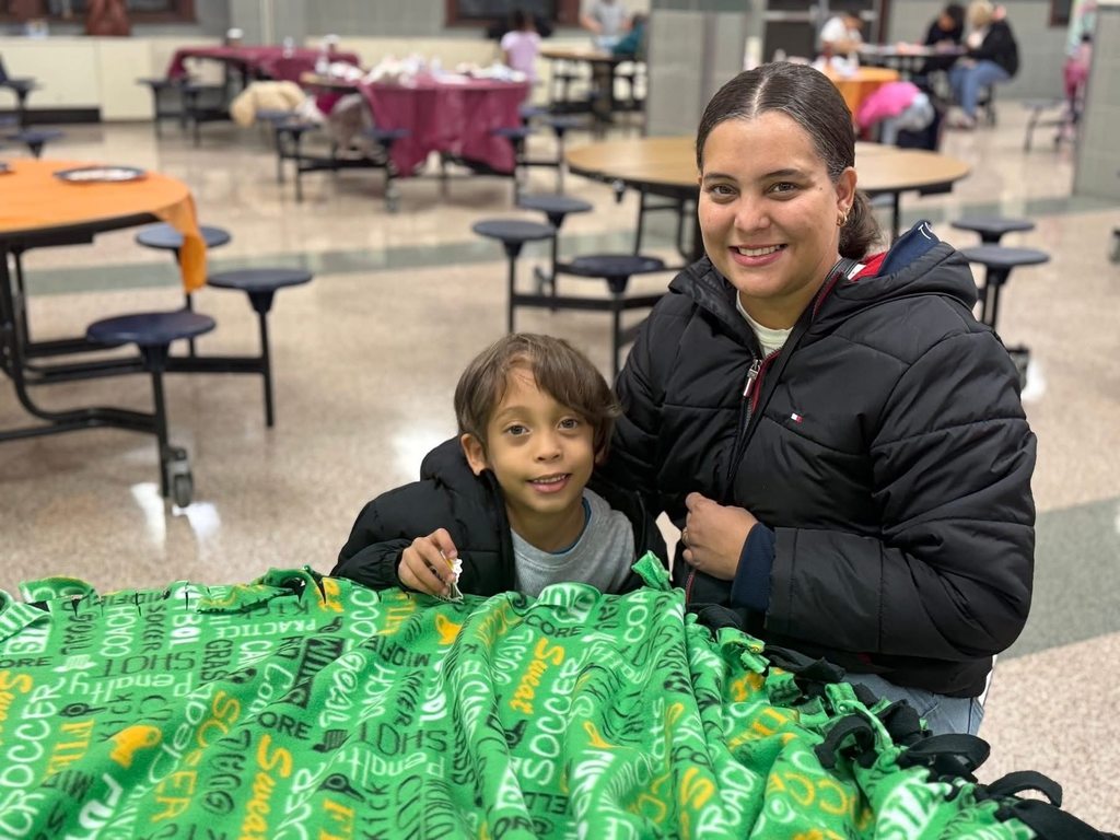 An adult woman and a child are sitting at a table, working on a colorful green blanket in a school cafeteria. Other people can be seen in the background.