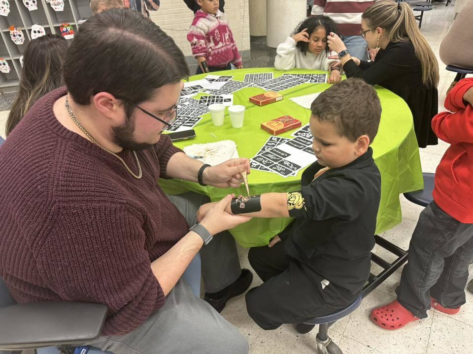 A district staff member is doing a henna painting on a young boy's arm who is sitting at a table in a school cafeteria. Other people can be seen in the background engaging in activities.