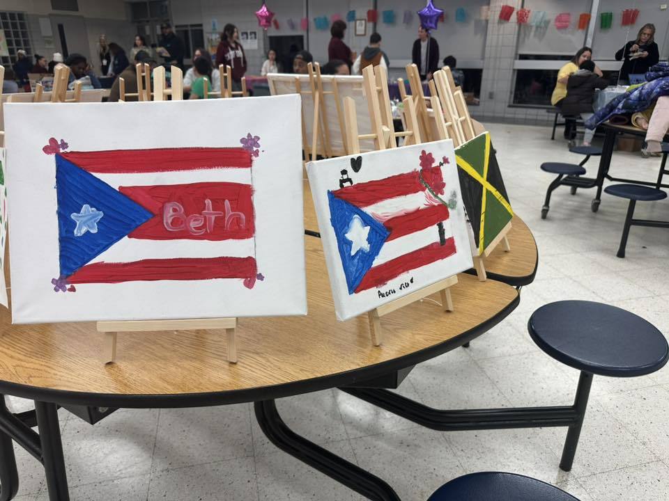 Art display with paintings of Puerto Rican flags on easels atop a round table in a school cafeteria. People and festive decorations can be seen in the background.