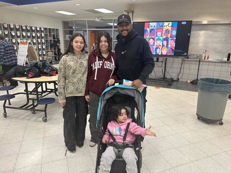 A family of four are posting together in a school cafeteria, with a stroller in front, amid tables and a colorful display that says "One World" in the background.
