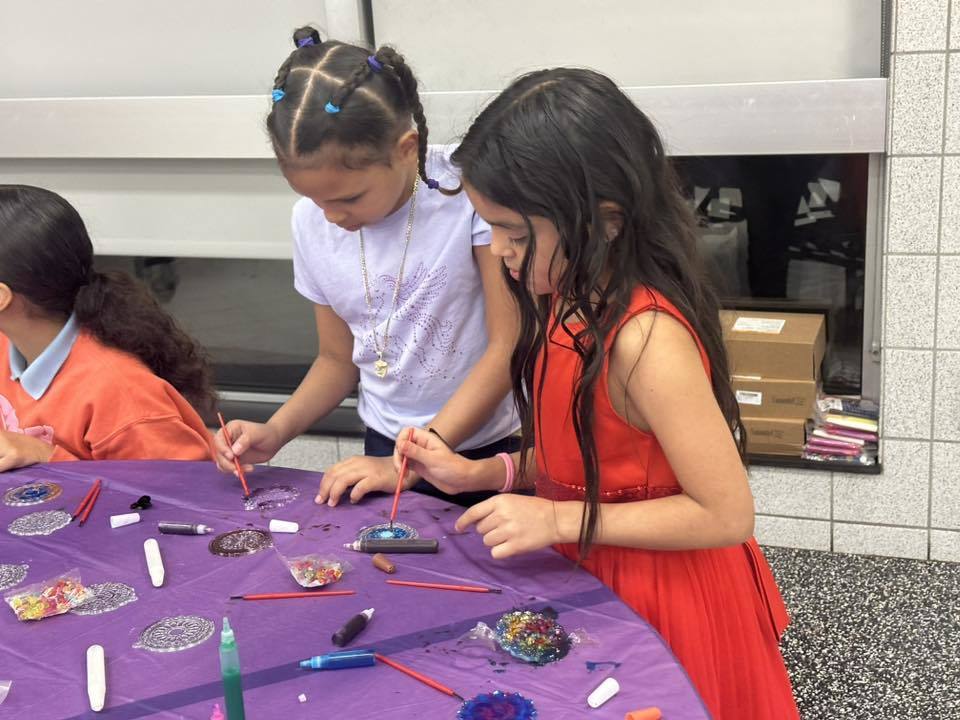 Two young girls are doing a craft activity together at a table in a school cafeteria. They are decorating small circular items with colorful materials. Another young girl can be seen nearby.
