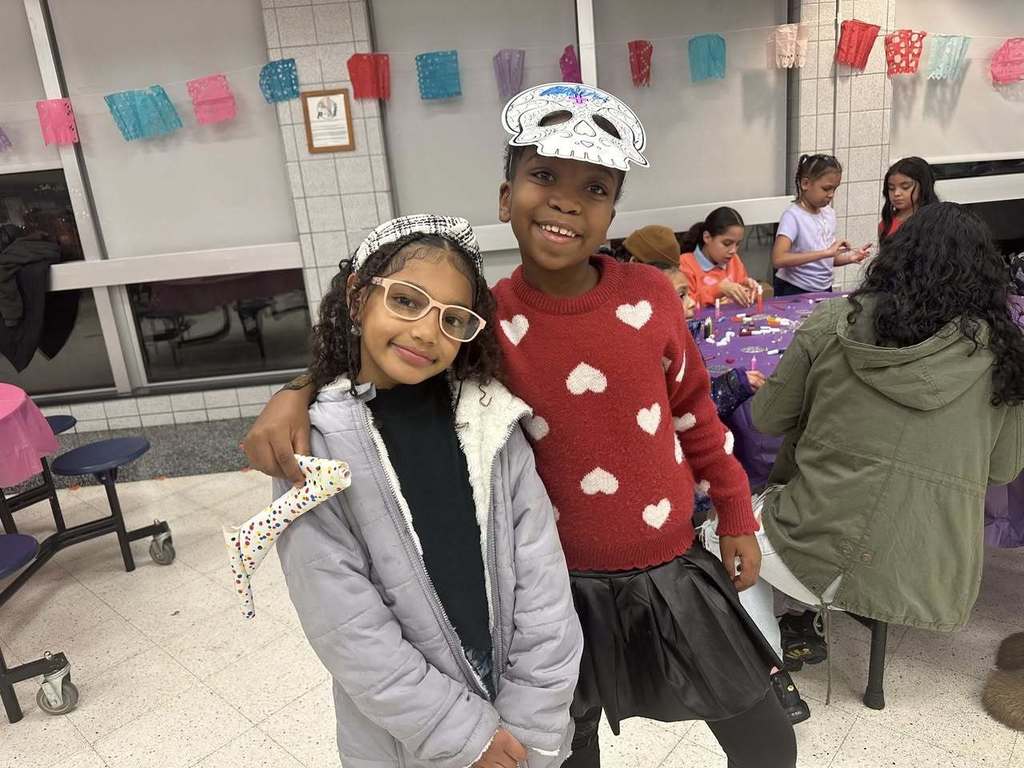 Two young girls are standing together in a school cafeteria, with one of the girls having her arm wrapped around the other girl. Other children can be seen doing crafts in the background. Colorful decor and paper crafts are hanging on the walls.