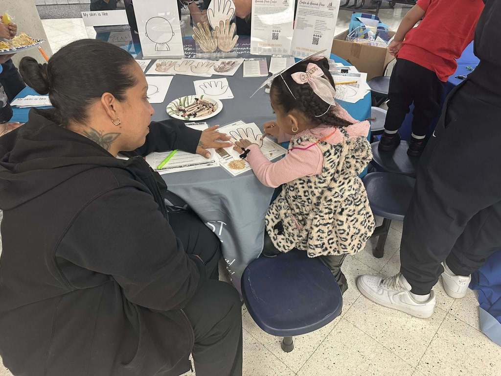 An adult is observing a young girl who is drawing on a piece of paper at a table in a school cafeteria. Other people can be seen nearby. 