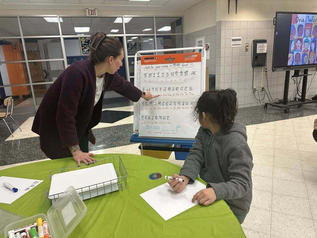 A district staff member is pointing at a whiteboard with Hindi script as a young girl is seated at a table with a marker in her hand in a school cafeteria.