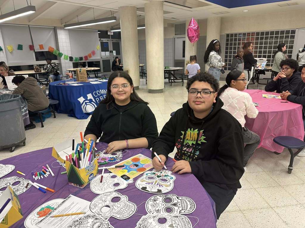 A teenage girl and boy are sitting at a table in a school cafeteria with coloring supplies and skull designs. The school cafeteria is decorated festively, with other students engaging in crafting activities nearby.