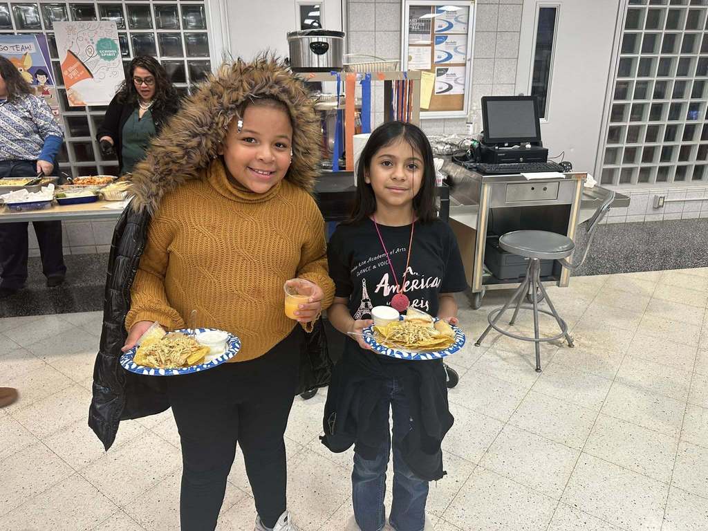 Two young girls are smiling and holding plates of food inside a school cafeteria. Other people and dishes of food can be seen in the background.