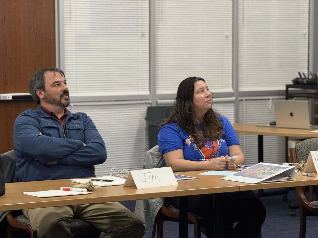 Two district staff members are sitting at a table in a conference room, attentively listening. Papers, pens, and name cards are on the table.