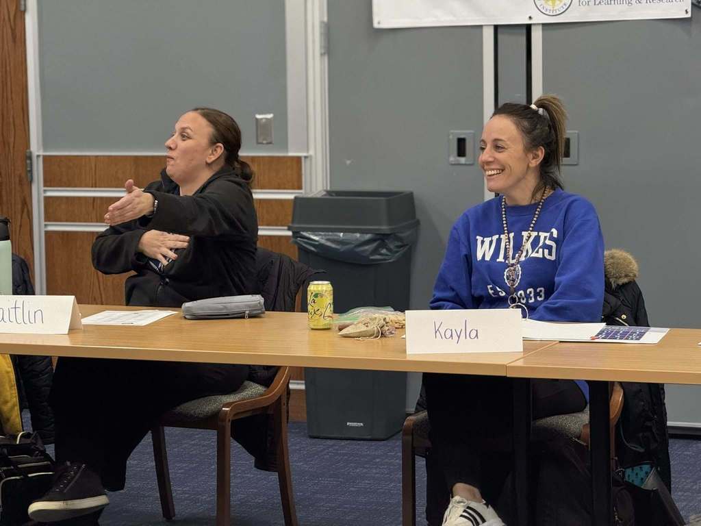 Two district staff members are sitting a table with name tags in a conference room. One of the woman is gesturing with her hand while she is speaking and the other woman is laughing.
