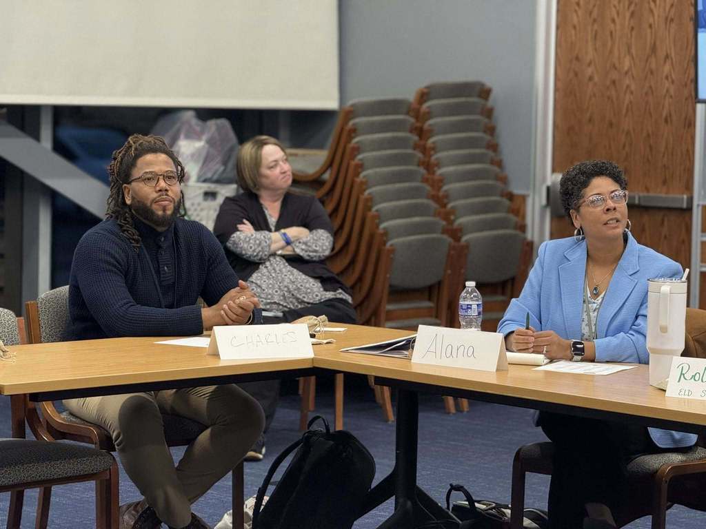 Two district staff members are sitting attentively at a table with name tags in a conference room. Another district staff member can be seen sitting next to two stacks of piled chairs in the background.