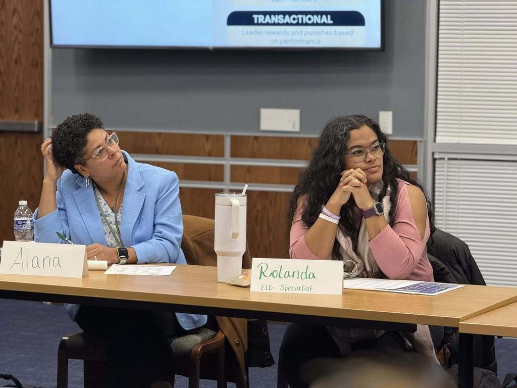 Two district staff members are sitting attentively at a table in a conference room. Name tags are in front of them.