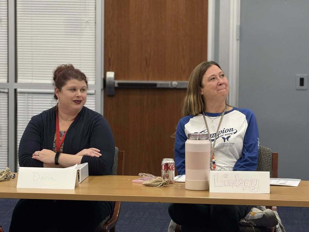 Two district staff members are sitting at a table in a conference room with name tags, attentively listening. Drinks are in front of them. 