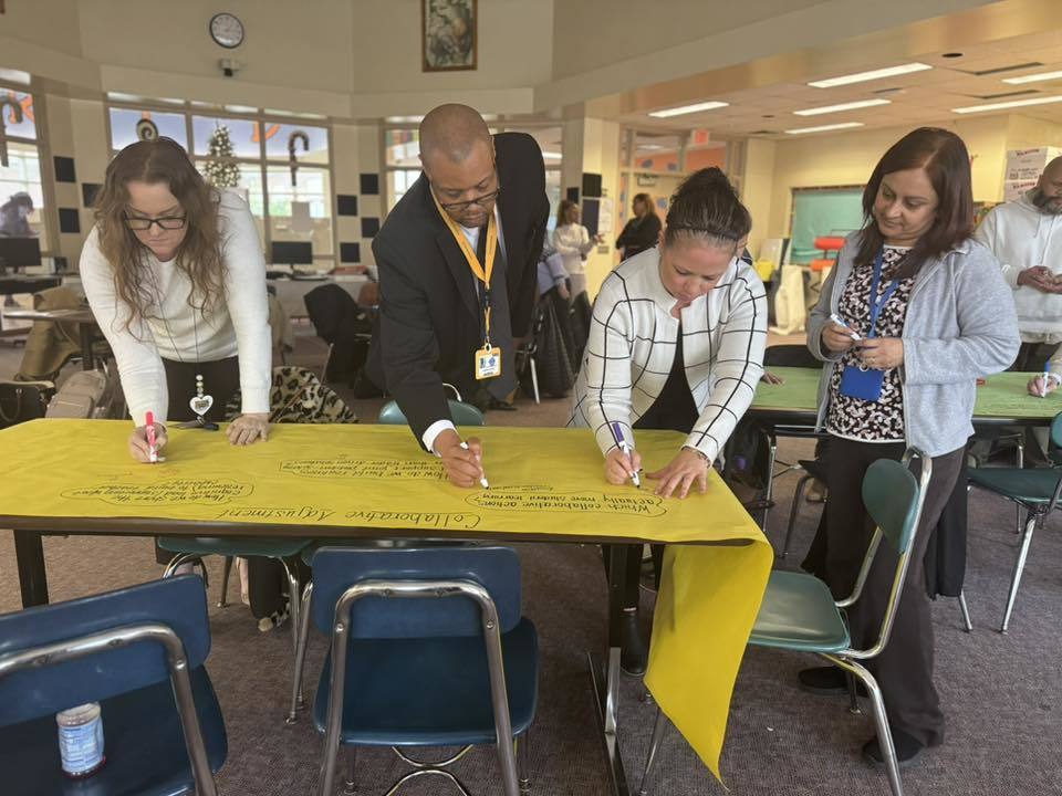 A group of four district staff members are collaboratively writing on a large yellow paper using markers in a school library. A box of markers are sitting on the table. Other people can be seen in the background.