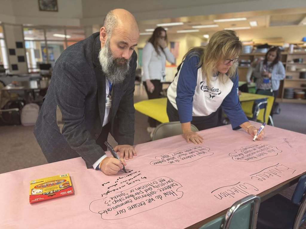 Two district staff members are collaboratively writing on a large pink paper with markers in a school library. A box of markers are sitting on the table. Other people can be seen in the background.