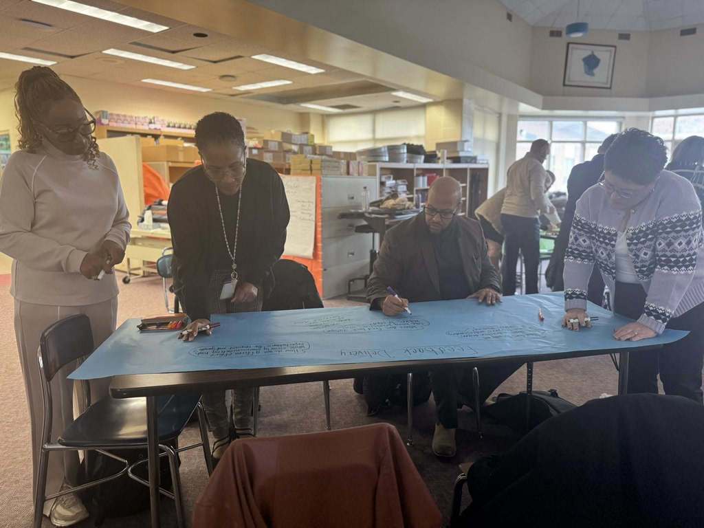 A group of four district staff members are collaboratively writing on a large blue paper using markers in a school library. A box of markers are sitting on the table. Other people can be seen in the background.