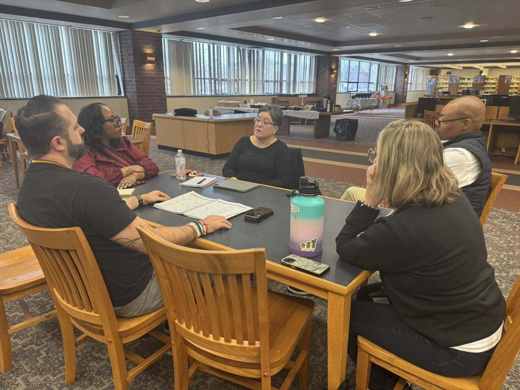 A group of five district staff members are engaging in a discussion at a table in a school library. Papers and water bottles can be seen on the table.