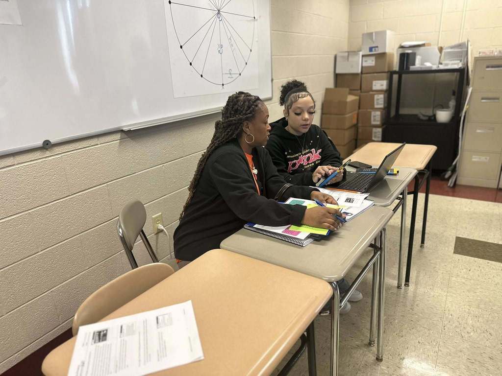A district staff member and a student are sitting at desks in a school classroom looking at a laptop, engaging in discussion.
