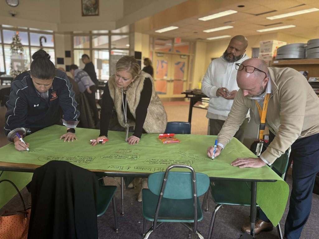 A group of four district staff members are collaboratively writing on a large green paper with markers in a school library. A box of markers are sitting on the table. Other people can be seen in the background.