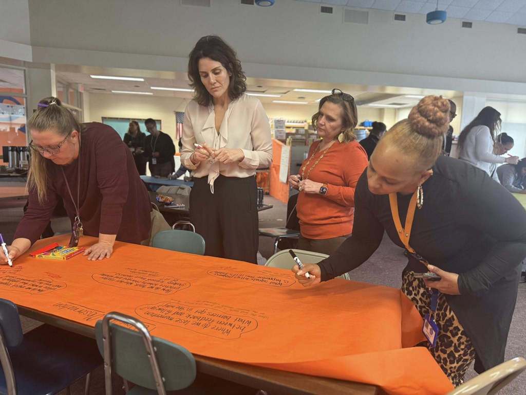 A group of four district staff members are collaboratively writing on a large orange paper using markers in a school library. A box of markers are sitting on the table. Other people can be seen in the background.