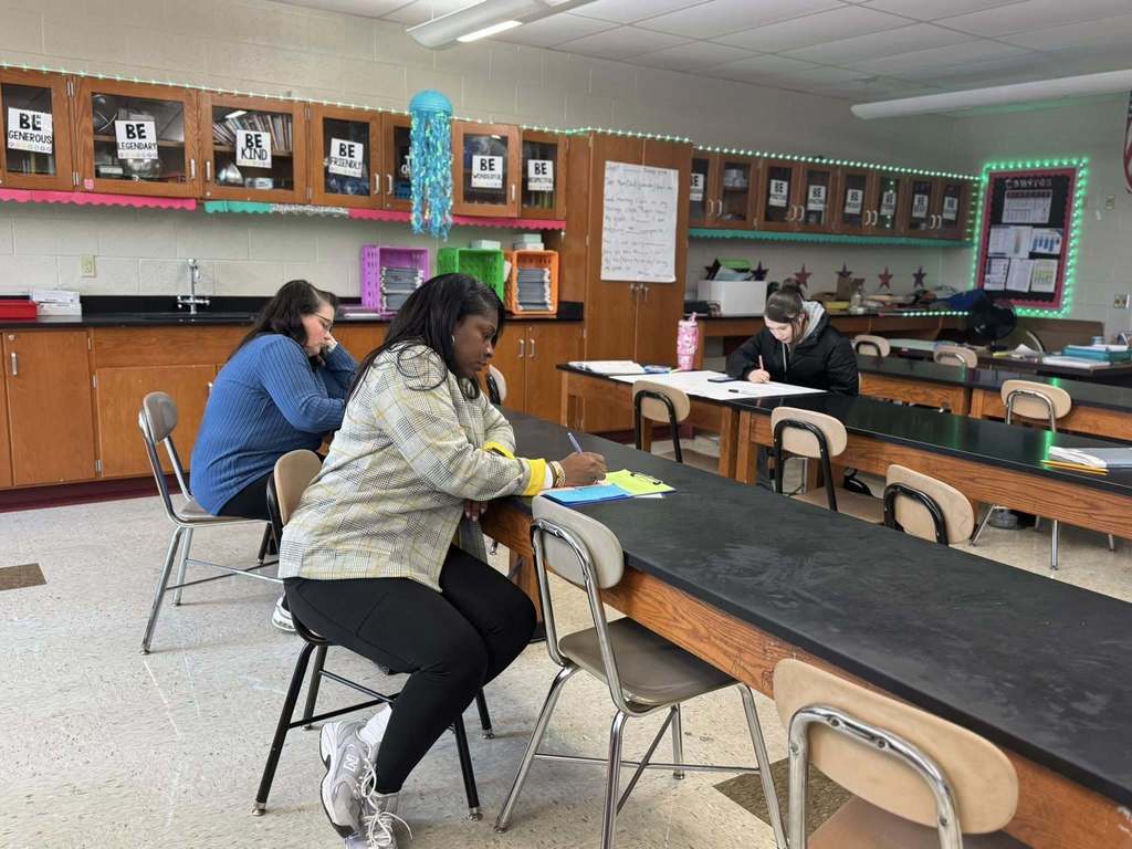 Two district staff members are seated at a lab table in a school classroom, writing on clipboards. A student can be seen sitting at another lab table nearby writing on a large poster.
