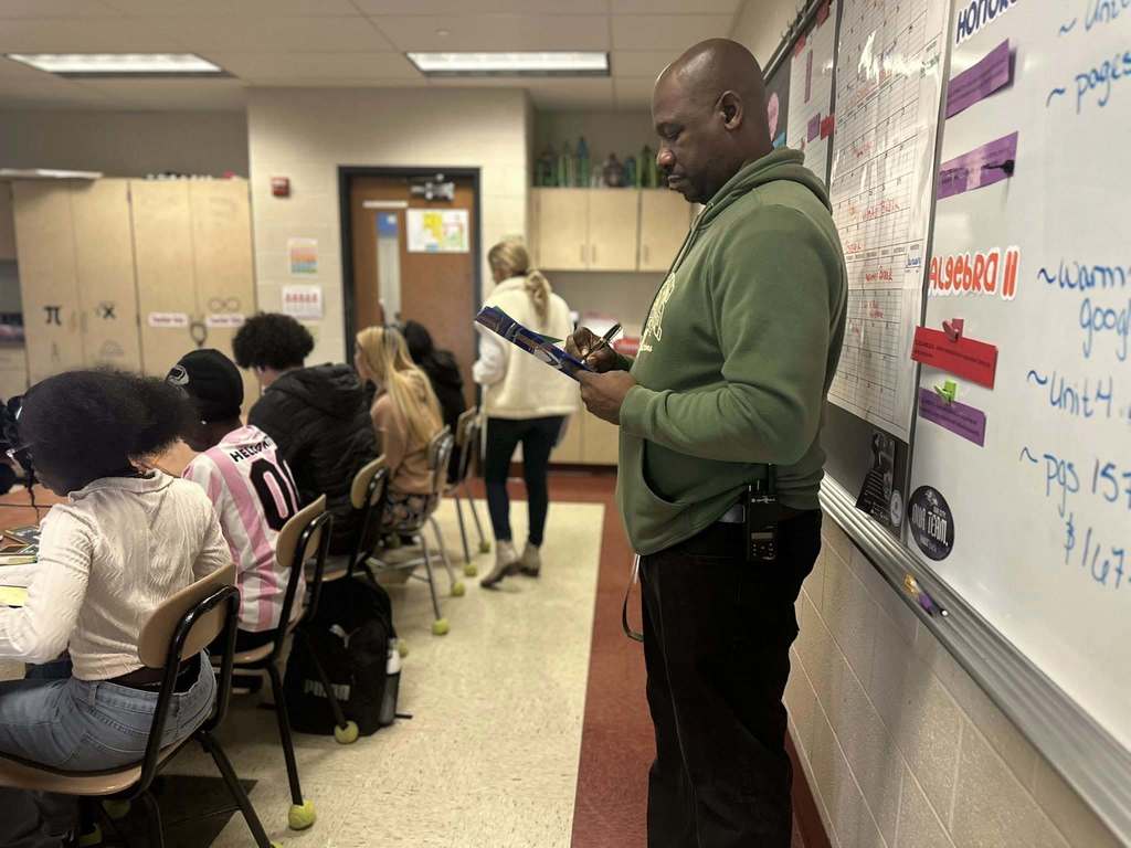 A district staff member is standing in front of a whiteboard writing on a clipboard in a school classroom. A teacher can be seen walking by a group of students who are seated at desks. 
