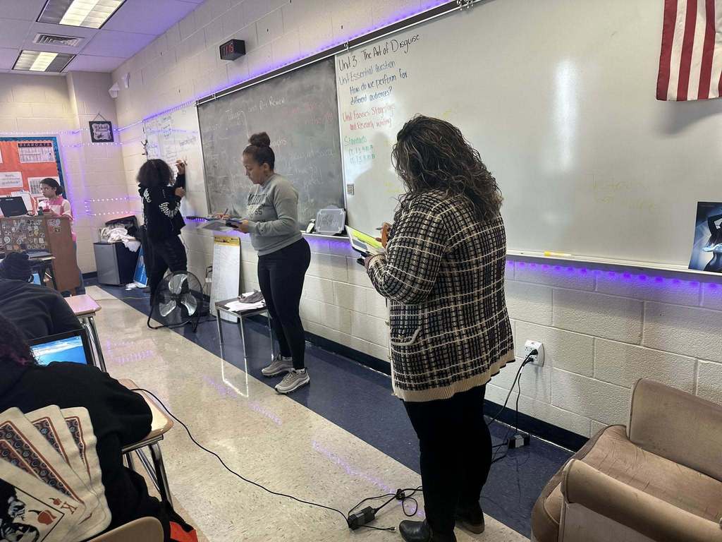 Two district staff. members are writing on clipboards in a school classroom. A student can be seen writing on a whiteboard nearby. Other students can be seen sitting at desks with laptops. 