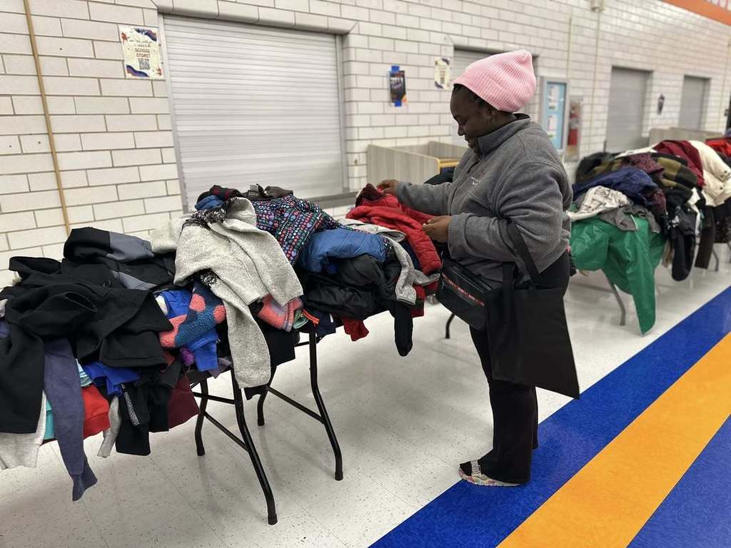 A woman is browsing a table stacked with winter coats in a school cafeteria.