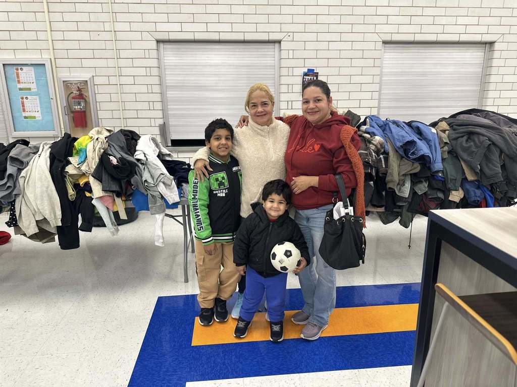 A family of four, including two adult women and two children, are standing in a school cafeteria with folded coats on a table in the background. One of the children is holding a soccer ball.